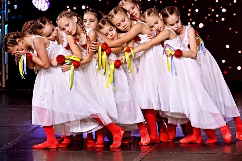 Girls in white dresses with blue and yellow ribbons and red boots performing an ensemble dance on stage