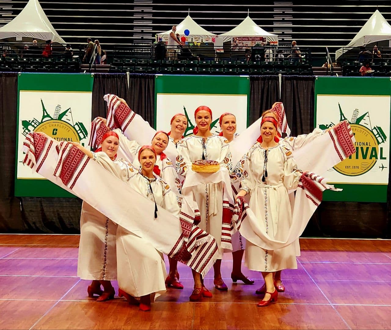 Adult folk ensemble in embroidered white dresses and red headscarves waving rushnyk towels and holding a korovai bread at a cultural festival